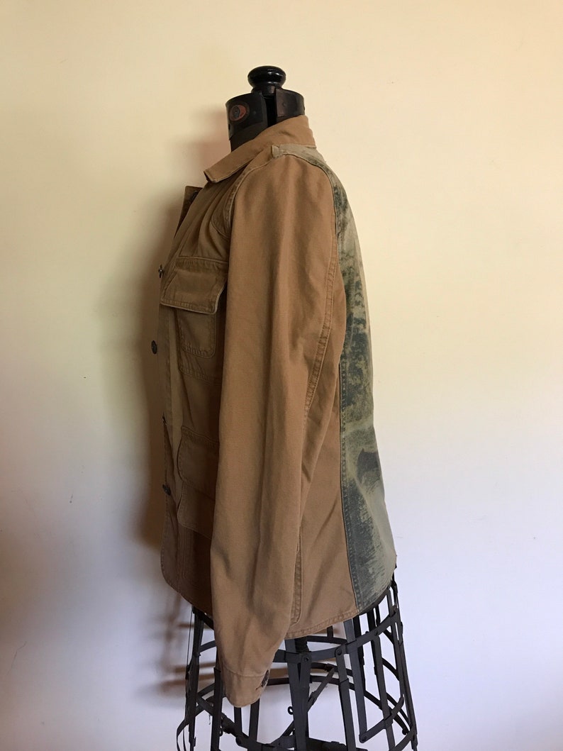May include: A brown utility jacket with a camouflage pattern on the back. The jacket has a button closure and two pockets on the front.
