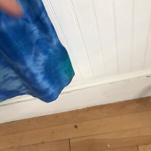 Puede incluir: Un primer plano de una cortina de color azul y verde con estampado tie-dye que cuelga frente a una pared blanca con lambrines. La cortina est&aacute; parcialmente retirada, dejando ver un suelo de madera.