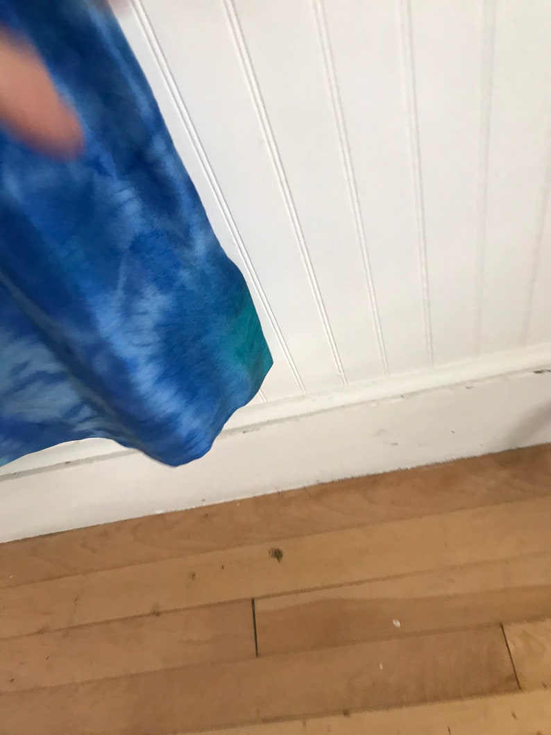 Puede incluir: Un primer plano de una cortina de color azul y verde con estampado tie-dye que cuelga frente a una pared blanca con lambrines. La cortina est&aacute; parcialmente retirada, dejando ver un suelo de madera.