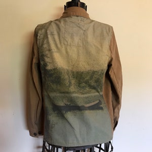 May include: A brown canvas jacket with a large green and blue print on the back. The print features a landscape scene with a boat on a lake.