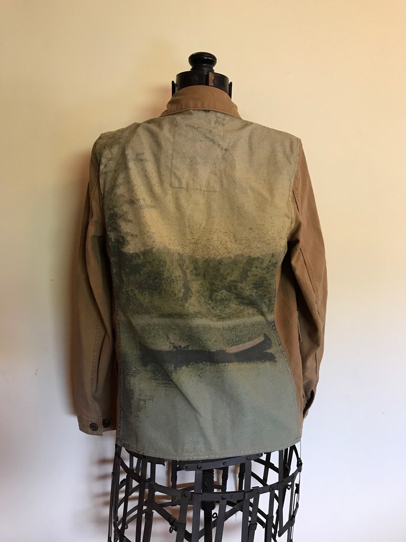 May include: A brown canvas jacket with a large green and blue print on the back. The print features a landscape scene with a boat on a lake.