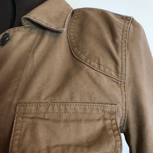 May include: A brown canvas jacket with a button-up front, a collar, and a large pocket on the left side. The jacket has a slightly distressed look.