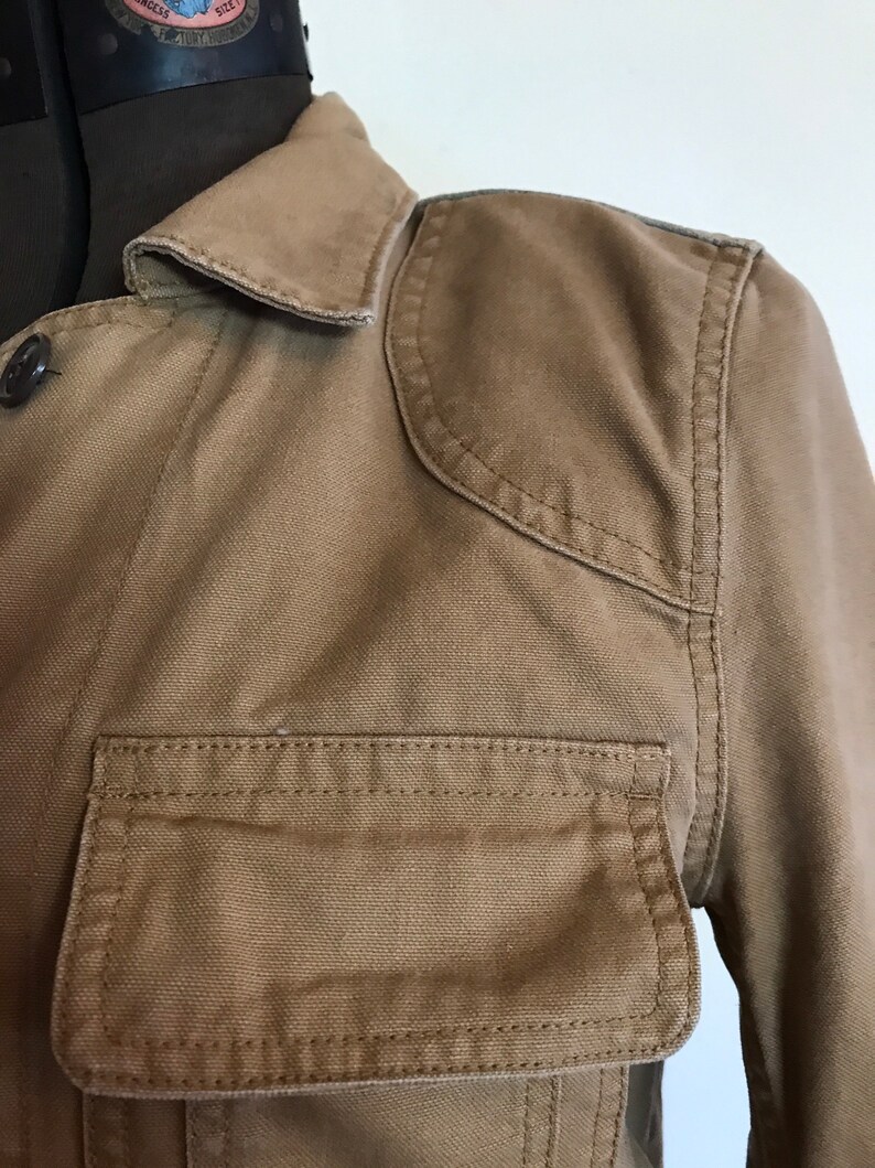 May include: A brown canvas jacket with a button-up front, a collar, and a large pocket on the left side. The jacket has a slightly distressed look.