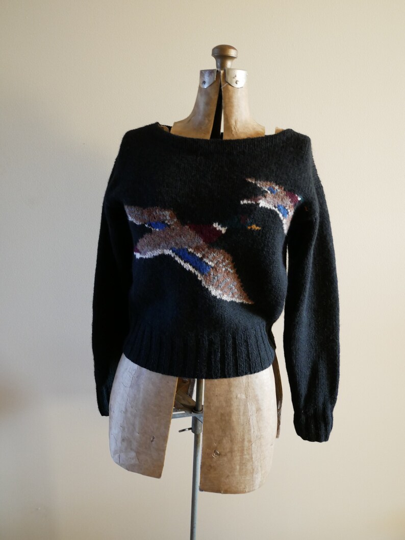 Pode incluir: Camisola preta com gola redonda e mangas compridas. A camisola apresenta um design colorido de p&aacute;ssaro no peito, com tons de castanho, azul e branco. A pe&ccedil;a de vestu&aacute;rio &eacute; exibida num manequim vintage.