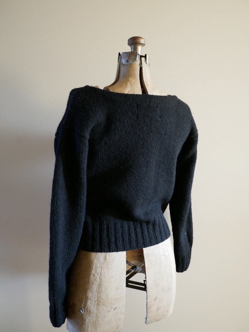 Pode incluir: Um su&eacute;ter preto com decote barco e mangas compridas, exibido em um manequim vintage. O su&eacute;ter tem bainha e punhos canelados. O fundo &eacute; de cor bege neutra. A pe&ccedil;a de roupa &eacute; de malha.