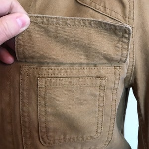 May include: Close-up of a brown canvas jacket with a large, double-stitched pocket on the left side.