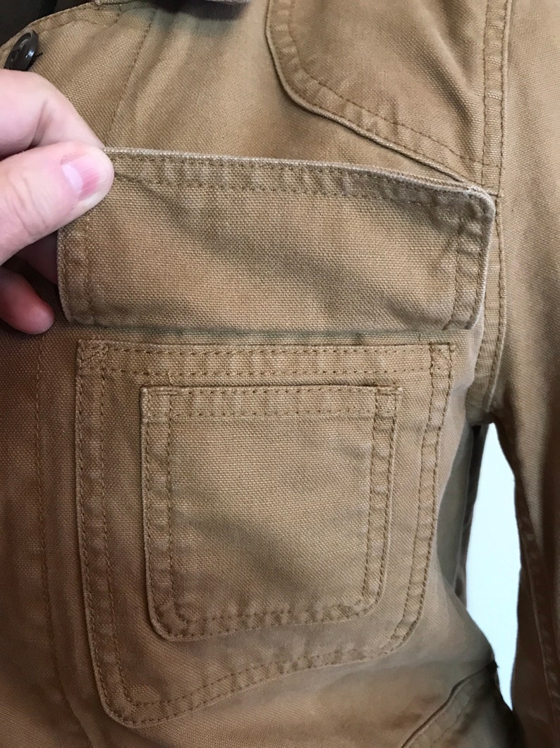 May include: Close-up of a brown canvas jacket with a large, double-stitched pocket on the left side.