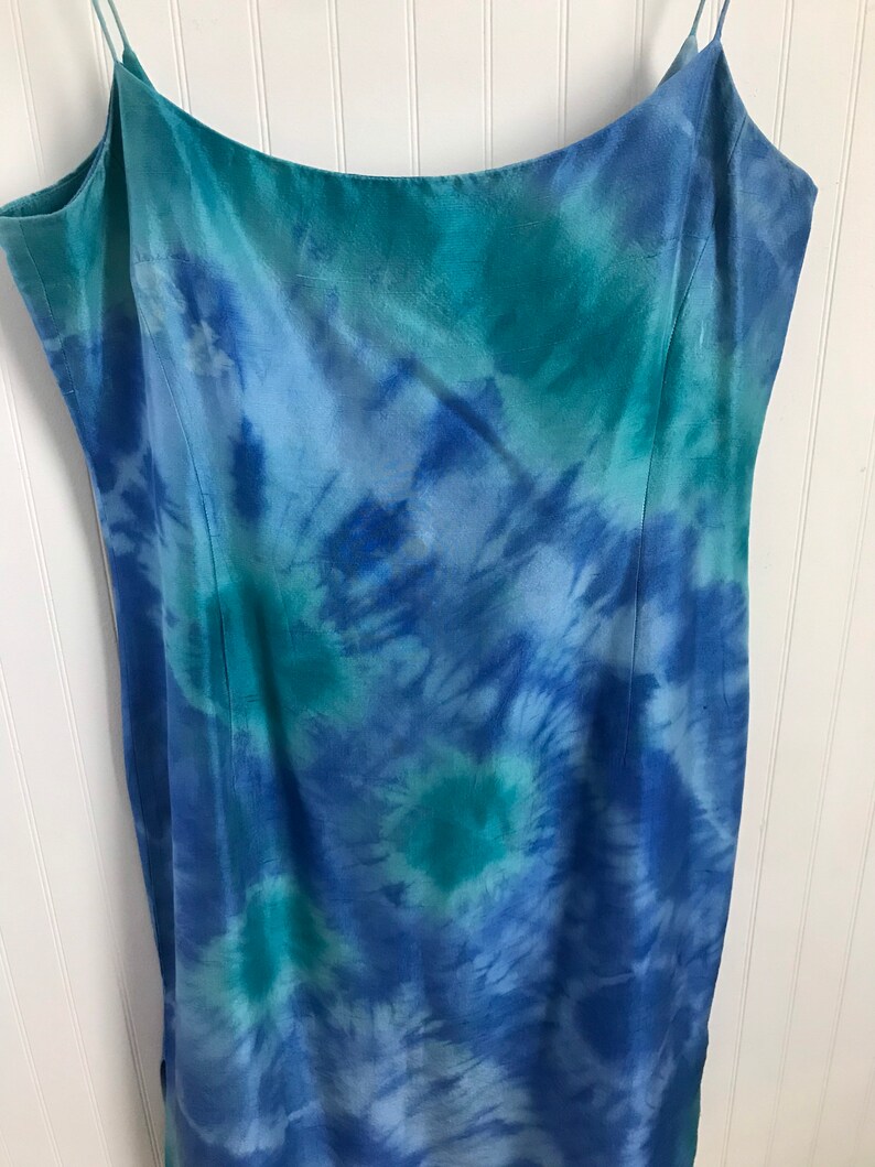 Puede incluir: Un vestido maxi de color azul y verde con estampado tie-dye y tirantes finos. El vestido est&aacute; hecho de una tela sedosa y tiene una silueta fluida.