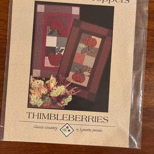May include: A package featuring two Harvest Table Toppers with apple and pumpkin designs. The toppers have a patchwork design with red, green, and brown colors. The package also includes the text "THIMBLEBERRIES" and "Classic Country by Lynette Jensen."