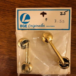Könnte beinhalten: Zwei goldfarbene Türknauf aus Metall mit rundem Design. Die Knäufe sind in einer durchsichtigen Plastiktüte verpackt, auf der eine weiße Karte mit der Aufschrift "BGE Originales Registered 1.55 25" angebracht ist.