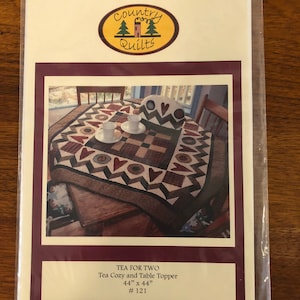 May include: A quilting pattern for a tea cozy and table topper. The pattern is called "Tea for Two" and is 44 inches by 44 inches. The pattern features a checkered design with hearts and circles in brown, red, and cream colors.