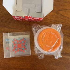 Puede incluir: Una caja abierta con un patrón rojo y blanco, un paquete de plástico transparente y una herramienta de costura naranja. El paquete contiene un patrón de estrella roja y azul. La herramienta naranja tiene las palabras "Jenny Makes Sewing Easy" impresas.