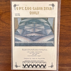 Pode incluir: Um molde de papel para um 6 Pt. Log Cabin Star Quilt de Vesta M. Ropp, datado de 1984. A capa apresenta uma fotografia de uma colcha com padrão de estrela azul e branco, com texto incluindo o título, LCC105, LCC106 e o endereço da empresa.