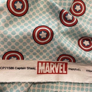 May include: Fabric featuring a pattern of Captain America's shield in red, white, and blue, set against a light blue polka dot background. The Marvel logo is visible on a white strip of fabric.