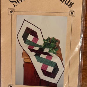 Op de afbeelding: Verpakking voor een naaipatroon genaamd "Ribbon Twist" van Stitchin' Friends. Het patroon toont een geometrisch ontwerp in wit, bordeauxrood, groen en roze. De verpakking heeft een beige achtergrond met de tekst "Stitchin' Friends".