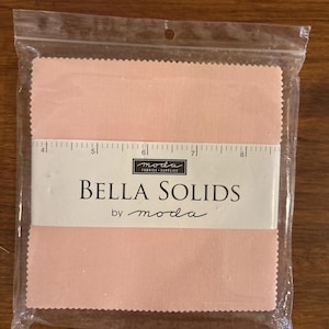 Puede incluir: Un paquete de cuadrados de tela Bella Solids en un tono rosa claro. La tela está empaquetada en una bolsa de plástico transparente. La etiqueta dice "Bella Solids by moda" e incluye una regla para medir. Los cuadrados de tela miden unos 20 cm de largo.