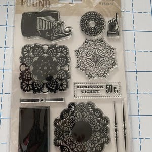 May include: Clear stamp set with 11 stamps. Includes a vintage camera, doily designs, a hand pointing up, a banner, and an admission ticket that reads "50c". The package reads "Lost & Found" and "My Mind's Eye".