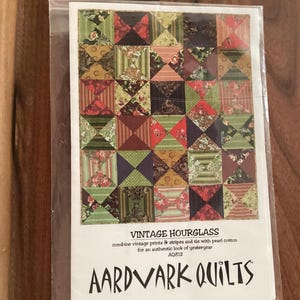 Op de afbeelding: Een pakket met een quiltpatroon getiteld "Vintage Hourglass". Het patroon toont een patchworkontwerp met vierkanten in verschillende kleuren en patronen, waaronder bloemen- en gestreepte stoffen. De tekst op de verpakking luidt "AARDVARK QUILTS."