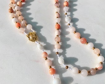 May include: A rosary featuring round beads in shades of peach, pink, and white, interspersed with small gold-tone metal spacer beads on a white cord. It includes a gold-tone metal centerpiece depicting a figure and a gold-tone crucifix.