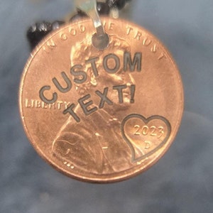 May include: A copper penny pendant with the text "CUSTOM TEXT!" engraved on it. The penny has a heart with the year 2023 engraved on it. The pendant is attached to a black beaded necklace.