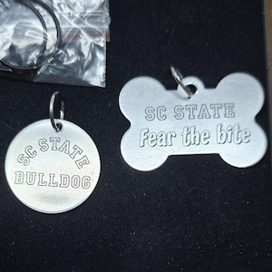 May include: A set of two silver dog tags in a cardboard box. One tag is round with "SC STATE BULLDOG" engraved. The other is bone-shaped with "SC STATE Fear the bite" engraved. Includes a small bag with a split ring.