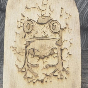May include: Wooden spatula with a frog design. The spatula is light brown with a frog carved into the wood. The handle has a series of carved footprints. The spatula is on a gray background.