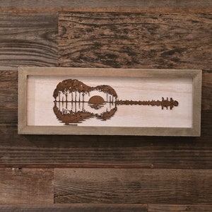 Puede incluir: Una pieza de arte de pared de madera enmarcada que presenta una silueta de una guitarra con una puesta de sol y &aacute;rboles reflejados en el cuerpo de la guitarra.