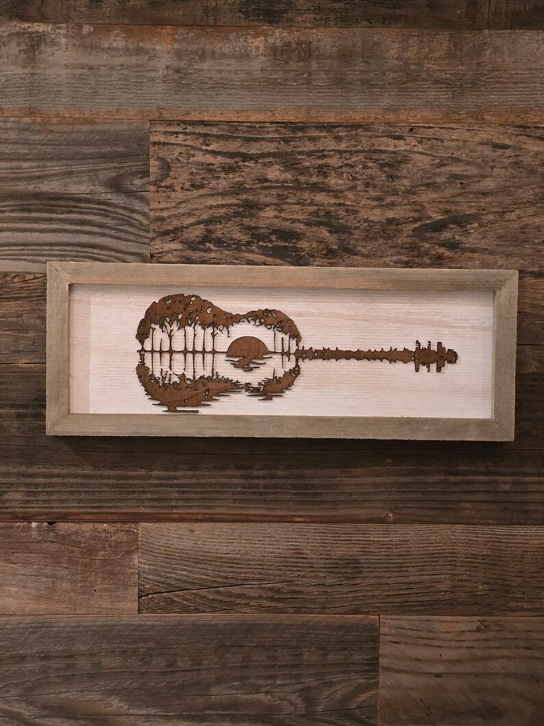 Puede incluir: Una pieza de arte de pared de madera enmarcada que presenta una silueta de una guitarra con una puesta de sol y &aacute;rboles reflejados en el cuerpo de la guitarra.
