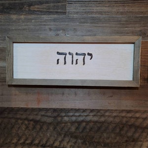 May include: A wooden sign with a light brown frame and a white background. The sign has the Hebrew word "קדושה" written in black.