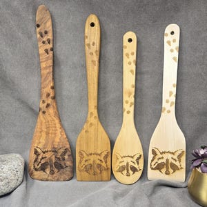 May include: Four wooden spatulas with raccoon faces and paw prints carved into them. The spatulas are different shades of brown and natural wood.
