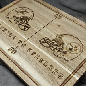 May include: A wooden cutting board with an engraved design of two football helmets, one for the Miami Dolphins and one for the Pittsburgh Steelers. The board has the words "Dolphins vs Steelers" and the scores "50" and "50" engraved on it.