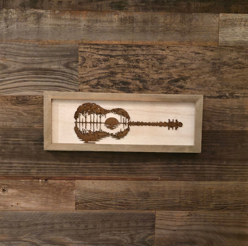 Puede incluir: Una pieza de arte de pared enmarcada en madera que presenta una silueta de una guitarra ac&uacute;stica con una puesta de sol y &aacute;rboles dentro del cuerpo de la guitarra.
