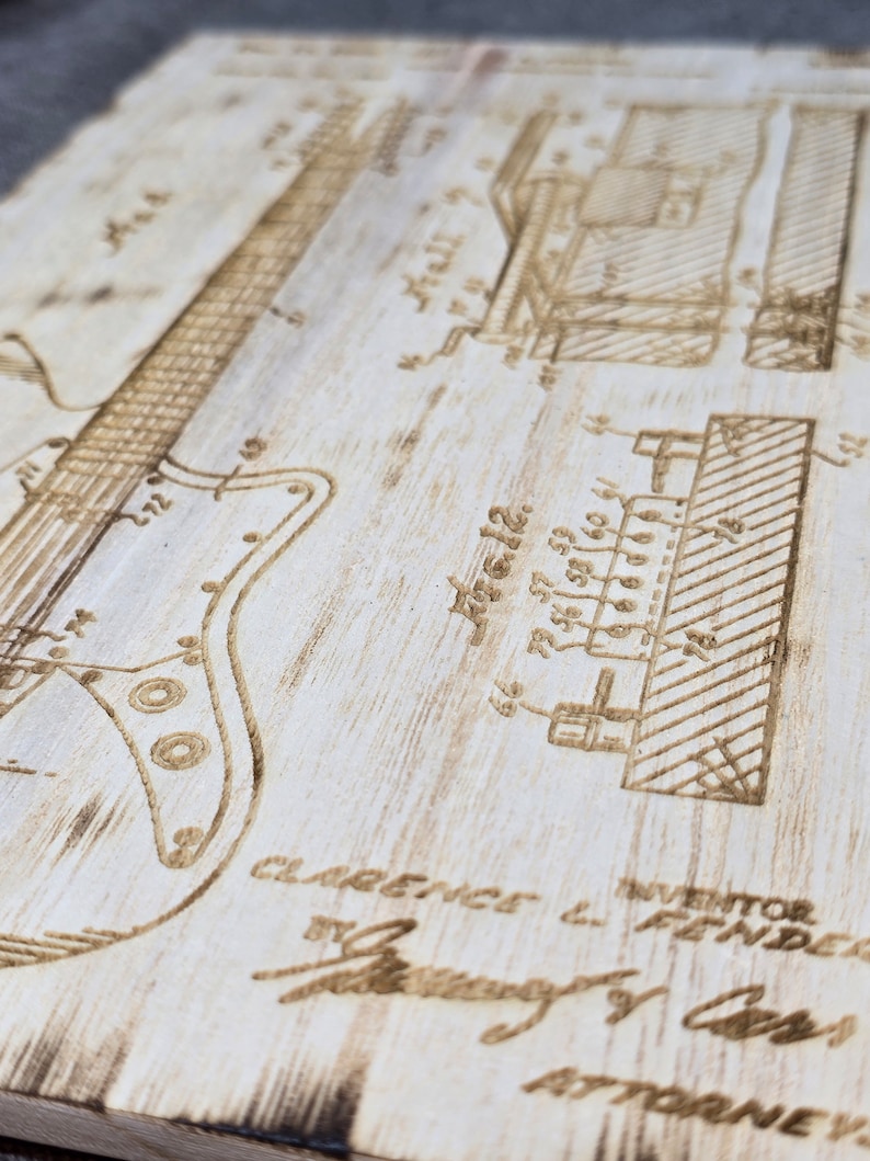 May include: A close-up of a wood-burned patent drawing of an electric guitar. The drawing includes the text "INVENTOR CLARENCE L. FENDER" and "ATTORNEY".