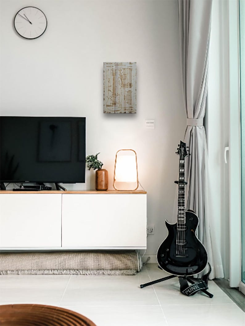 May include: A white wooden sign with a brown vintage guitar blueprint design. The sign is hanging on a white wall in a living room setting. A black electric guitar is on a stand in the foreground.