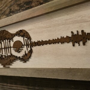 Puede incluir: Una pieza de arte de pared enmarcada en madera con una silueta marr&oacute;n de una guitarra con una puesta de sol y &aacute;rboles dentro del cuerpo de la guitarra.