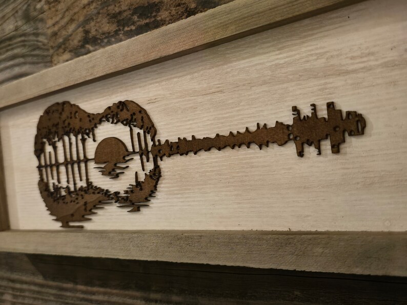 Puede incluir: Una pieza de arte de pared enmarcada en madera con una silueta marr&oacute;n de una guitarra con una puesta de sol y &aacute;rboles dentro del cuerpo de la guitarra.