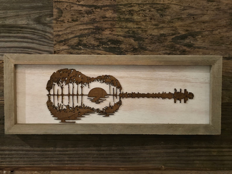 Puede incluir: Una pieza de arte de pared enmarcada en madera con una silueta marr&oacute;n de una guitarra con una escena de puesta de sol dentro del cuerpo de la guitarra.
