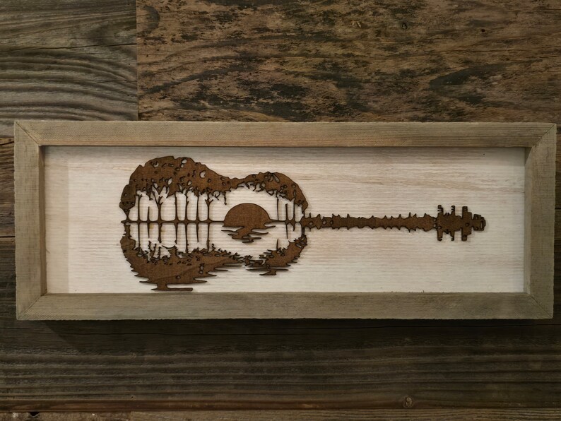Puede incluir: Una pieza de arte de pared enmarcada en madera con una silueta de una guitarra con una puesta de sol y &aacute;rboles dentro del cuerpo de la guitarra.