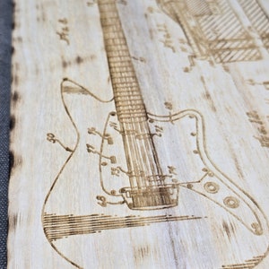 May include: A wood-burned design of an electric guitar with detailed line drawings of the body, neck, and strings. The design is on a light wood background.