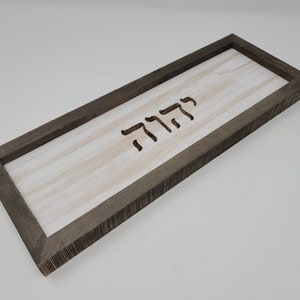 May include: A rectangular wooden sign with a gray frame and a white background. The word "שלום" is carved into the wood in Hebrew.