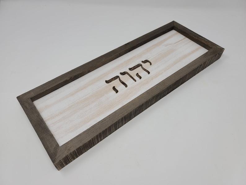 May include: A rectangular wooden sign with a gray frame and a white background. The word "שלום" is carved into the wood in Hebrew.