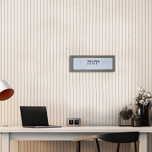 May include: A framed print with the Hebrew word "Chazak" in black lettering on a white background. The frame is silver and the print is hanging on a white wall.