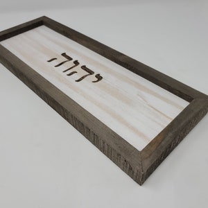 May include: A rectangular wooden sign with a white background and a dark brown frame. The sign has the Hebrew word "Shalom" carved into the wood.