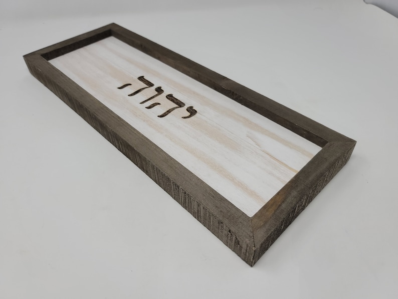 May include: A rectangular wooden sign with a white background and a dark brown frame. The sign has the Hebrew word "Shalom" carved into the wood.