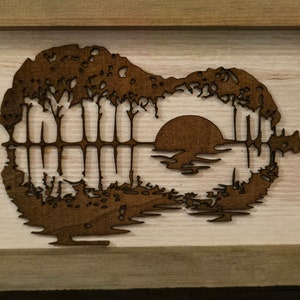 Puede incluir: Una pieza de arte de pared de madera con la silueta de una guitarra con una puesta de sol y &aacute;rboles reflejados en el cuerpo de la guitarra.