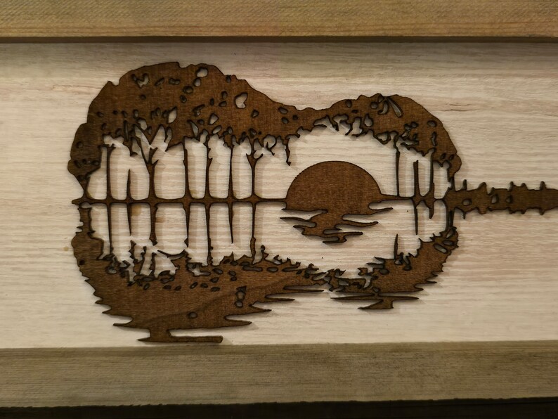 Puede incluir: Una pieza de arte de pared de madera con la silueta de una guitarra con una puesta de sol y &aacute;rboles reflejados en el cuerpo de la guitarra.