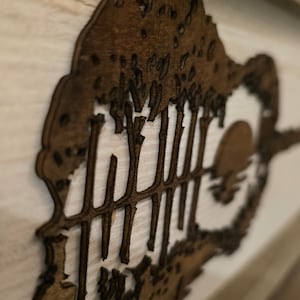 Puede incluir: Una pieza de arte de pared de madera que representa una silueta de una guitarra con &aacute;rboles en el fondo. La guitarra est&aacute; recortada en la madera y los &aacute;rboles est&aacute;n superpuestos detr&aacute;s de ella. La pieza est&aacute; pintada en un color marr&oacute;n oscuro.