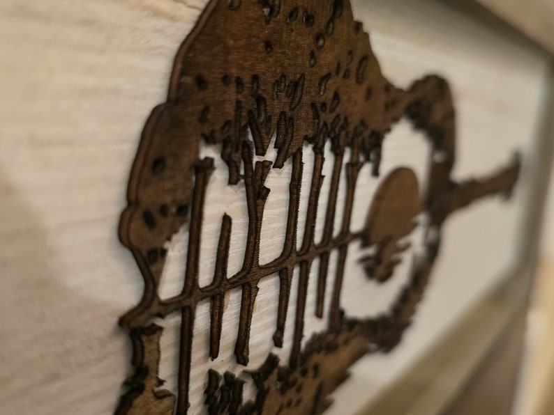 Puede incluir: Una pieza de arte de pared de madera que representa una silueta de una guitarra con &aacute;rboles en el fondo. La guitarra est&aacute; recortada en la madera y los &aacute;rboles est&aacute;n superpuestos detr&aacute;s de ella. La pieza est&aacute; pintada en un color marr&oacute;n oscuro.