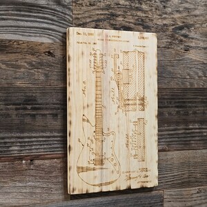 May include: A wood-burned image of a guitar patent drawing. The patent is for a "Bridge Construction for Guitars" and was filed on 24 February 1965, and granted on 13 December 1966. The patent number is 3,290,980. The patent drawing shows a detailed view of the guitar's bridge and the text "C.L. Fender" is visible.