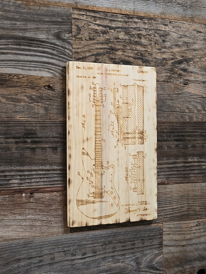 May include: A wood-burned image of a guitar patent drawing. The patent is for a "Bridge Construction for Guitars" and was filed on 24 February 1965, and granted on 13 December 1966. The patent number is 3,290,980. The patent drawing shows a detailed view of the guitar's bridge and the text "C.L. Fender" is visible.